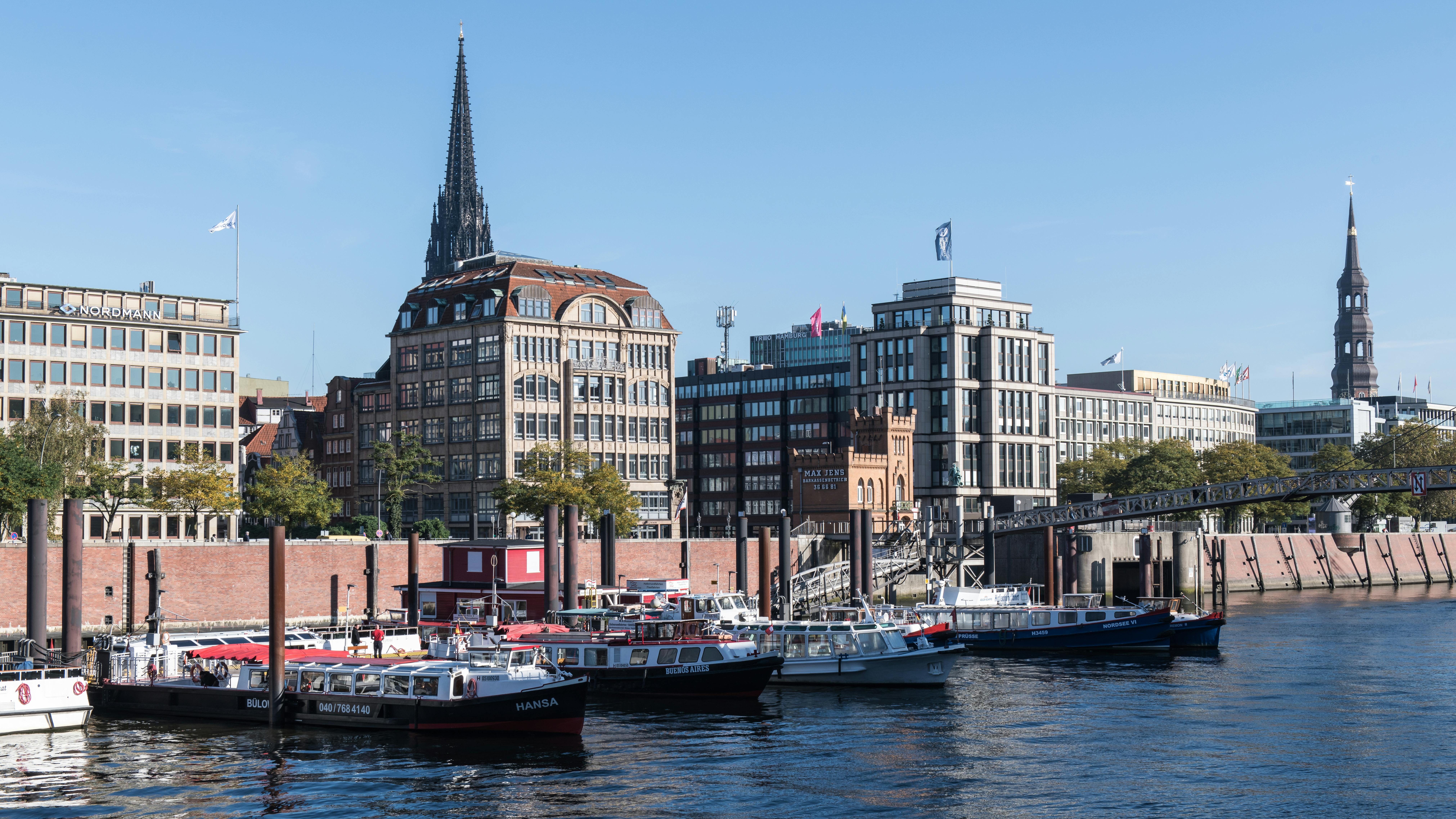 Hamburg waterfront skyline with historic church towers, modern buildings, and docked boats along the river under a clear blue sky.